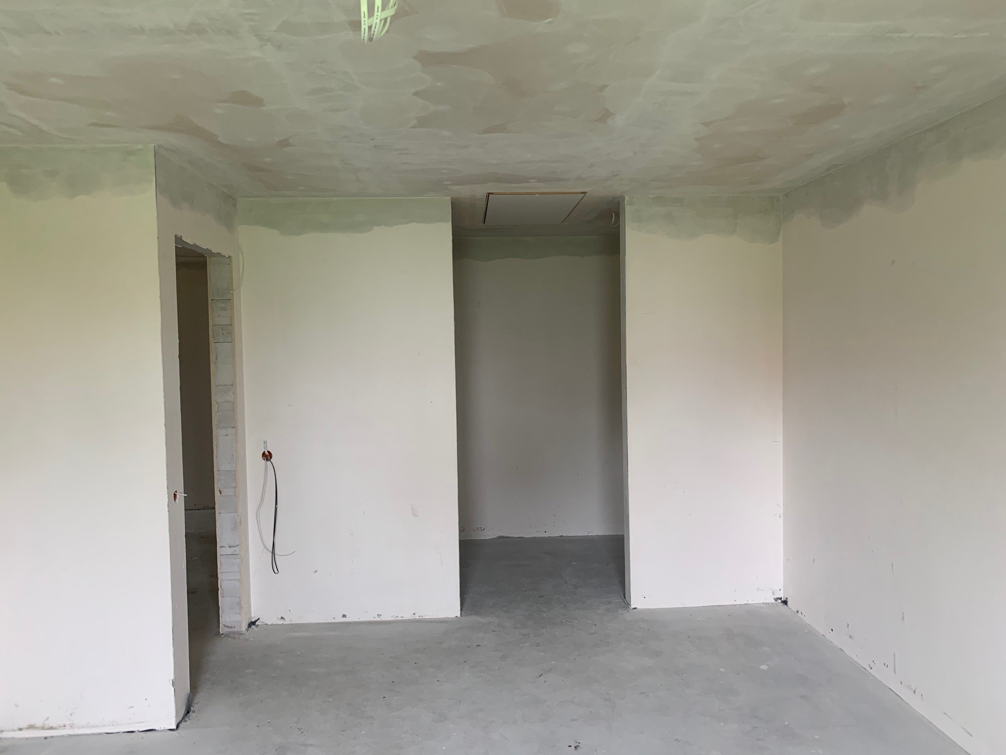 Wnętrze w trakcie prac wykończeniowych: gładzie na ścianach i suficie, surowa podłoga, widoczne otwory drzwiowe i instalacja elektryczna.