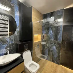 Nowoczesna łazienka po remoncie: kabina prysznicowa ze szklaną ścianą, umywalka na szafce, toaleta podwieszana, płytki imitujące marmur i drewno.