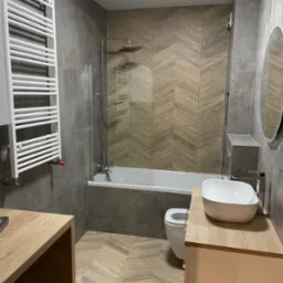 Nowoczesna łazienka z szarymi płytkami i drewnopodobną mozaiką na ścianie, biała umywalka nablatowa i grzejnik drabinkowy.