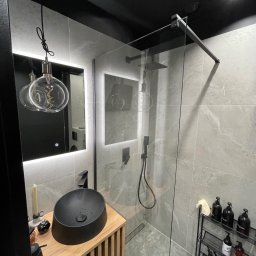 Wojciech Siemaszko - Nowoczesna łazienka z kabiną prysznicową typu walk-in, umywalką nablatową w kolorze czarnym i podświetlanym lustrem. Aranżacja w szarościach, z akcentami drewna.