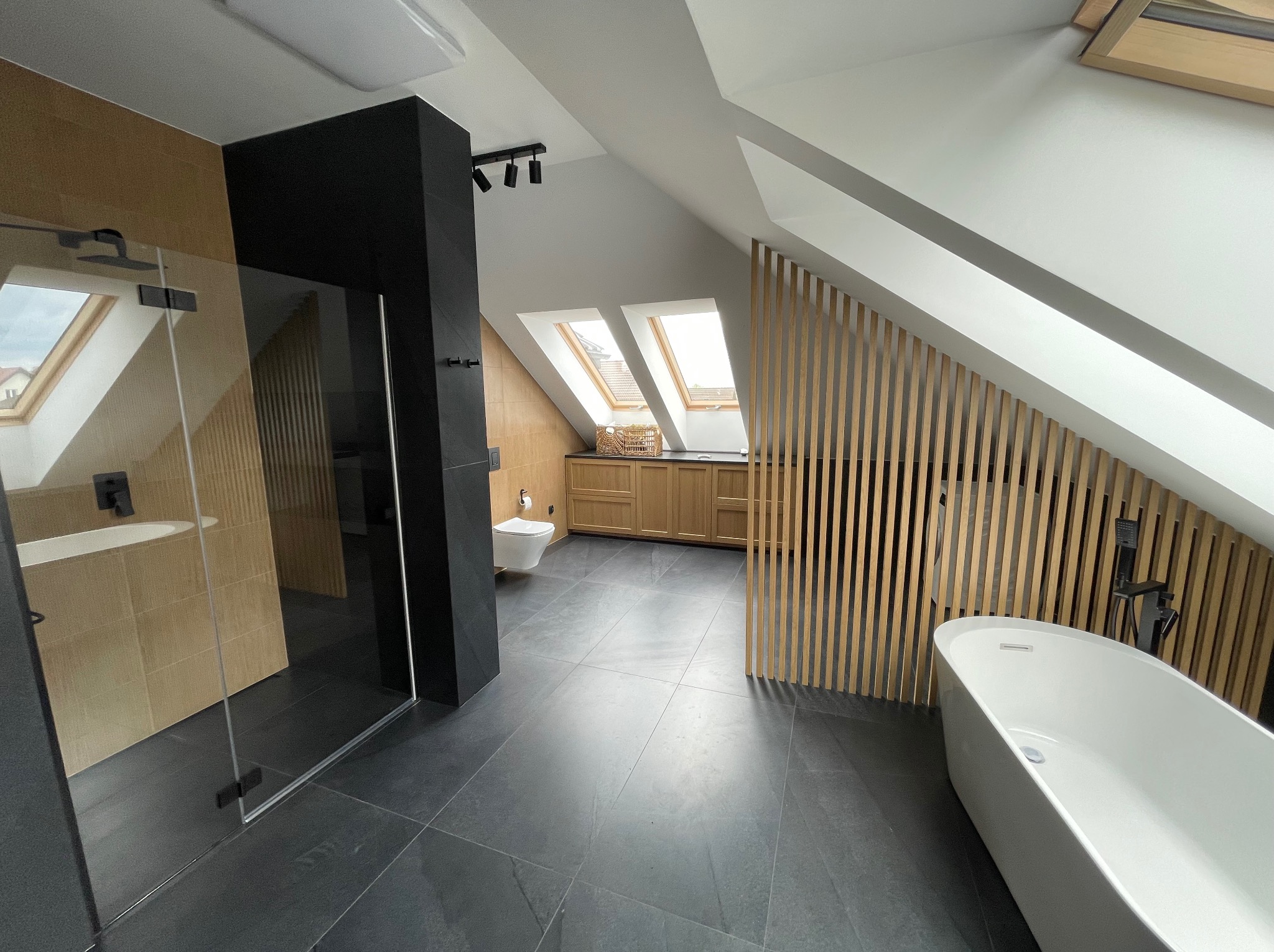Nowoczesna łazienka na poddaszu z kabiną prysznicową, wolnostojącą wanną i drewnianymi lamelami. Uwagę przykuwają okna dachowe w skośnej ścianie.