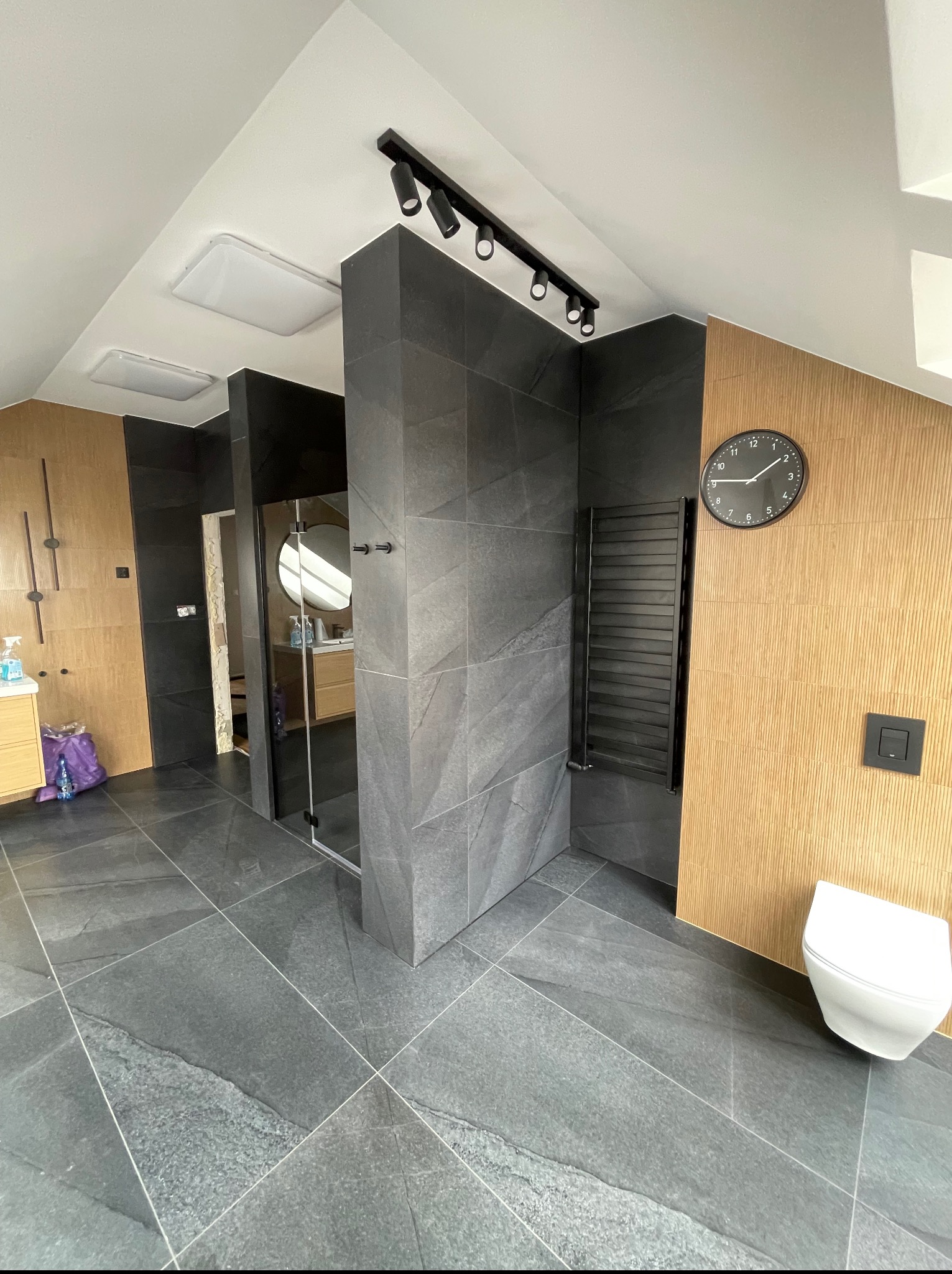 Wykończona łazienka na poddaszu z szarą, dużą płytką podłogową i kabiną prysznicową typu walk-in. Na ścianie drewnopodobna okładzina i zegar.