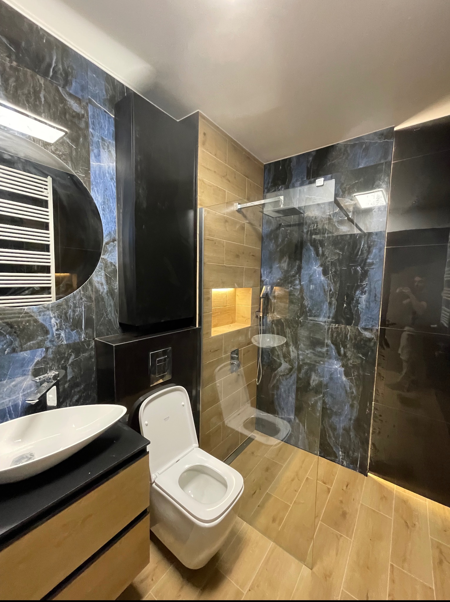 Nowoczesna łazienka po remoncie: kabina prysznicowa ze szklaną ścianą, umywalka na szafce, toaleta podwieszana, płytki imitujące marmur i drewno.