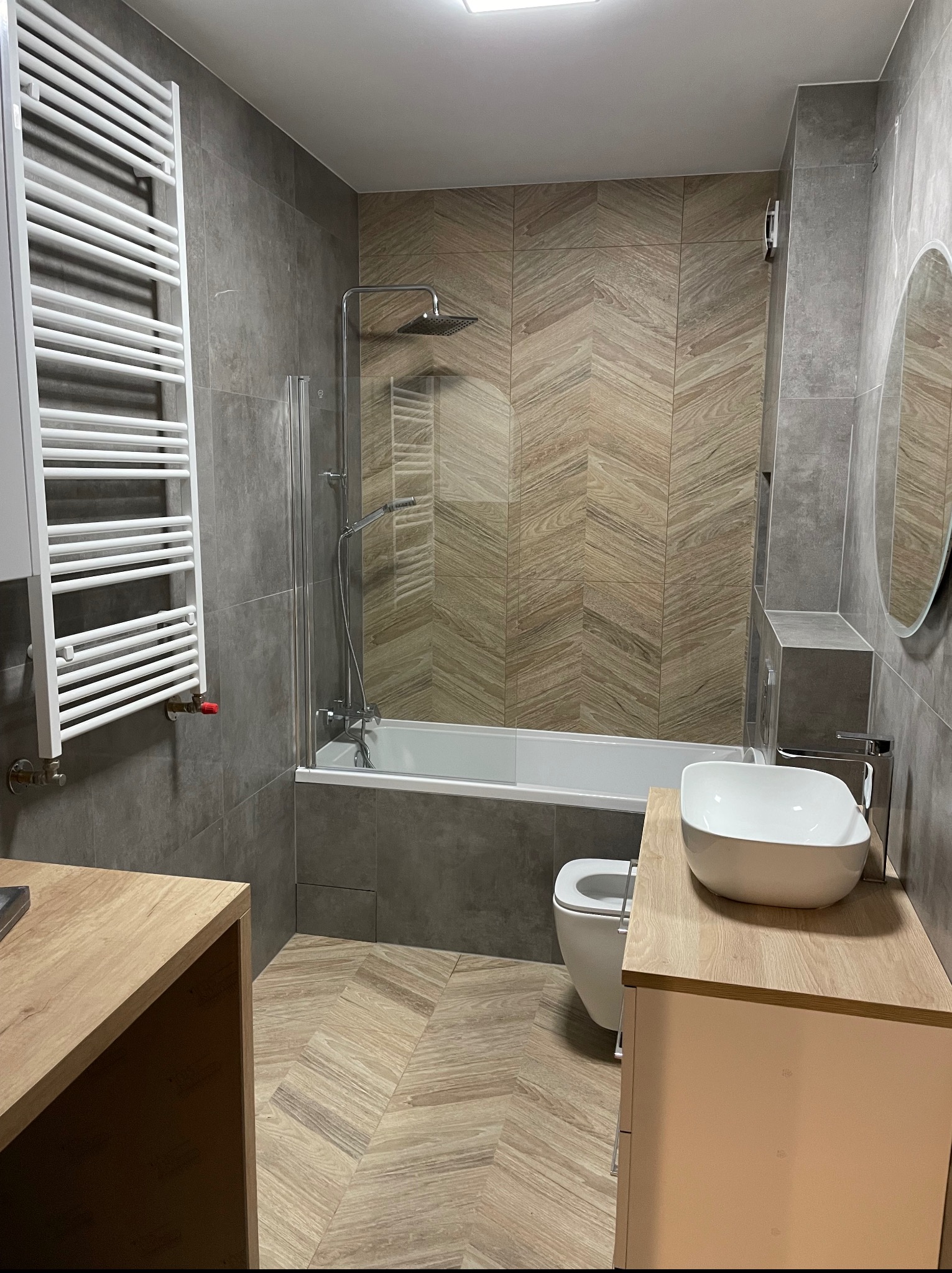 Nowoczesna łazienka z szarymi płytkami i drewnopodobną mozaiką na ścianie, biała umywalka nablatowa i grzejnik drabinkowy.