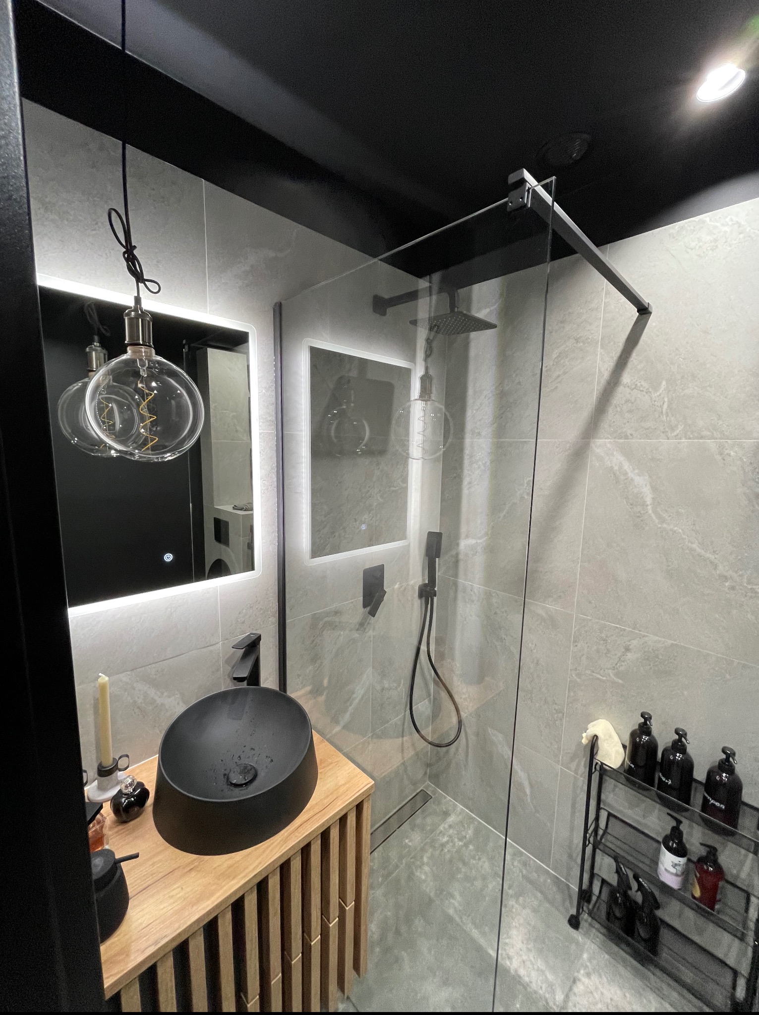 Nowoczesna łazienka z kabiną prysznicową typu walk-in, umywalką nablatową w kolorze czarnym i podświetlanym lustrem. Aranżacja w szarościach, z akcentami drewna.
