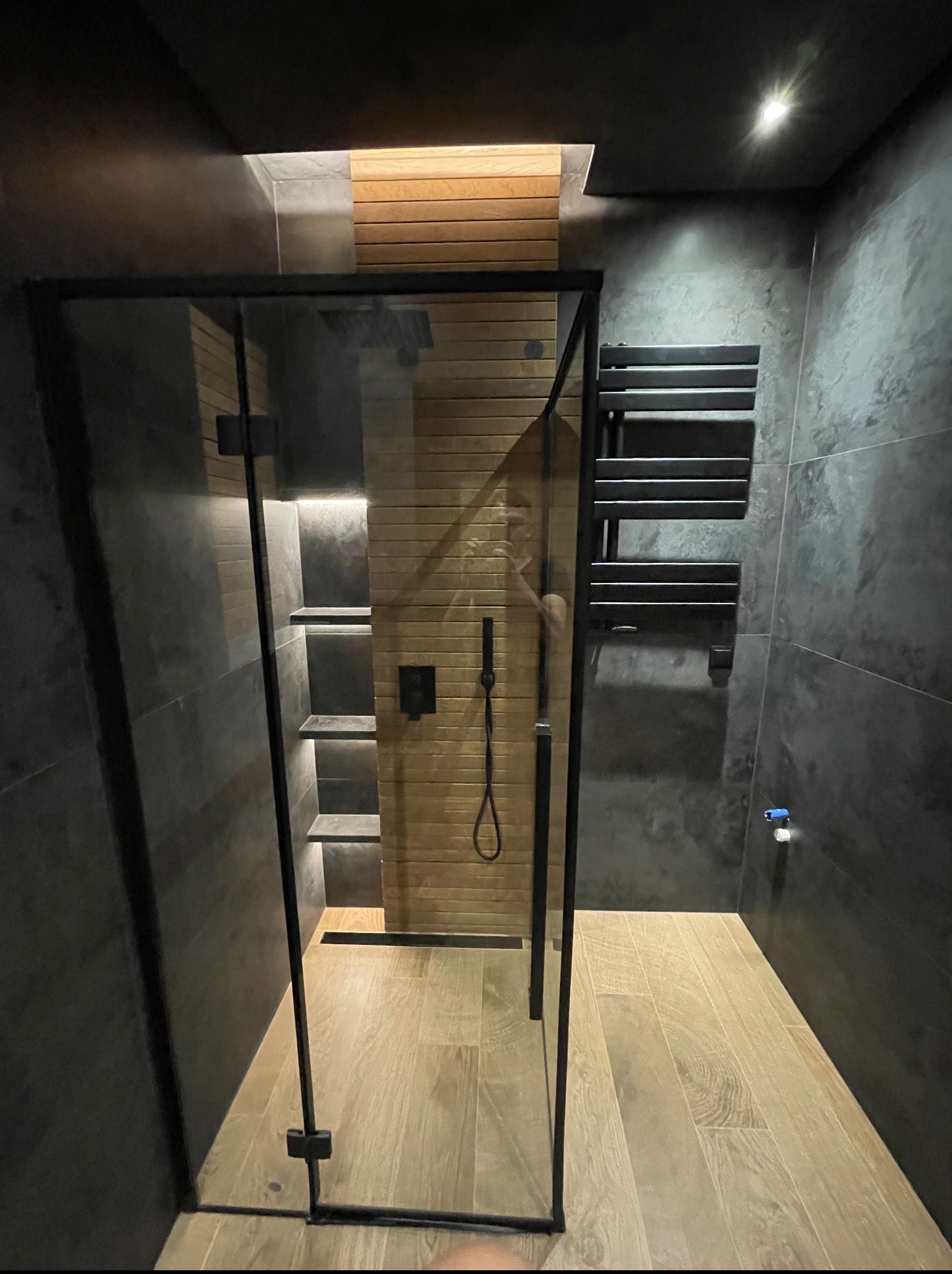 Nowoczesna łazienka z kabiną prysznicową walk-in, drewnianymi akcentami i grafitowymi płytkami. Minimalistyczny design, oświetlenie LED i grzejnik drabinkowy.