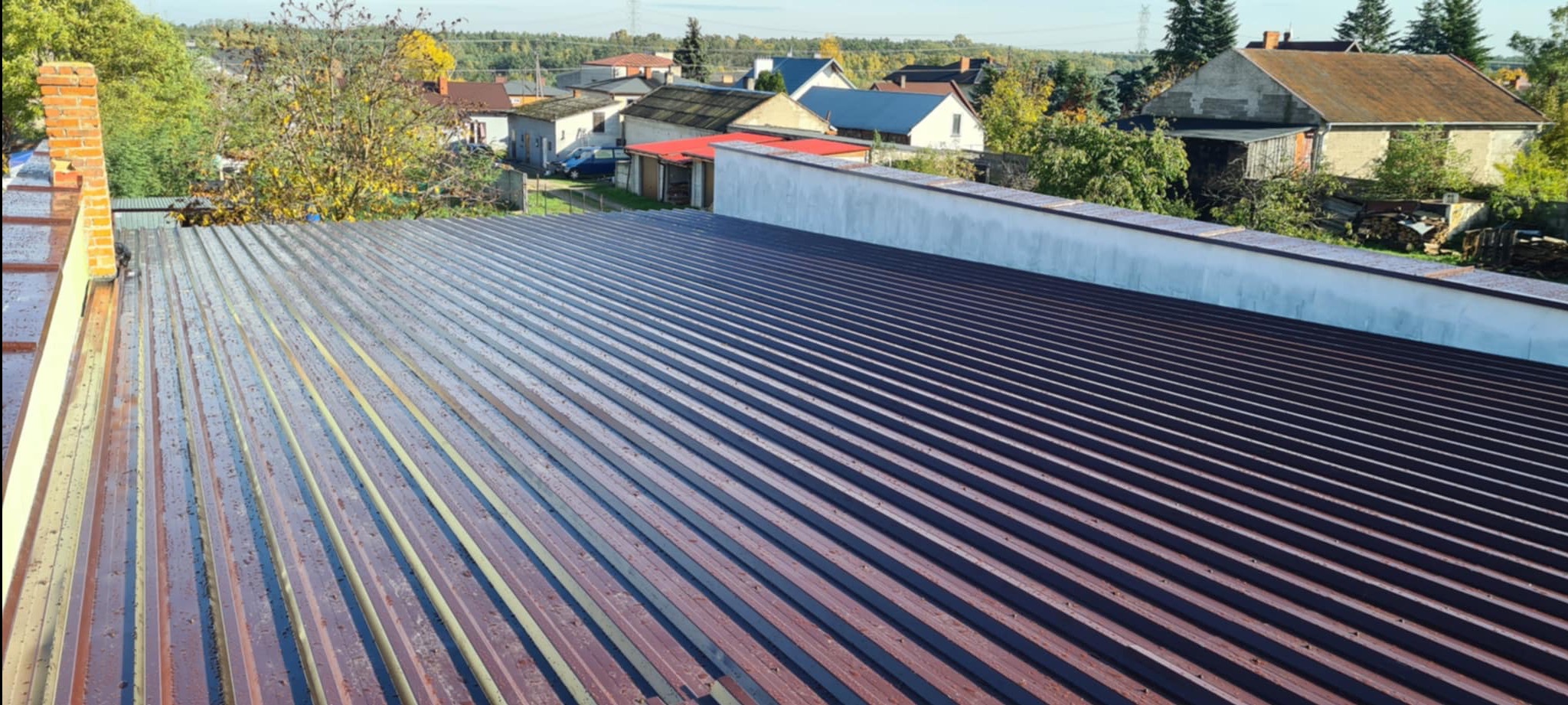 Nowy, brązowy dach z blachy trapezowej, widok z góry na tle zabudowań i drzew. Widoczne regularne rowki i mocowania blachy. Dach świeżo po montażu, w słoneczny dzień.