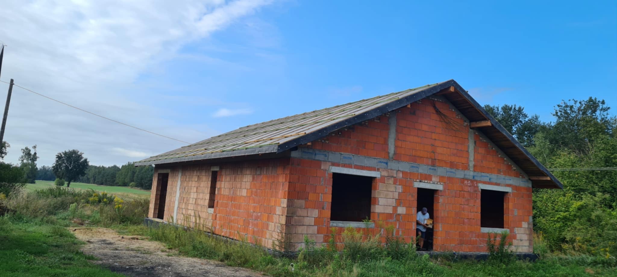 Niedokończony budynek z czerwonej cegły z częściowo położonym dachem, widoczne elementy więźby. W tle błękitne niebo i zieleń drzew.