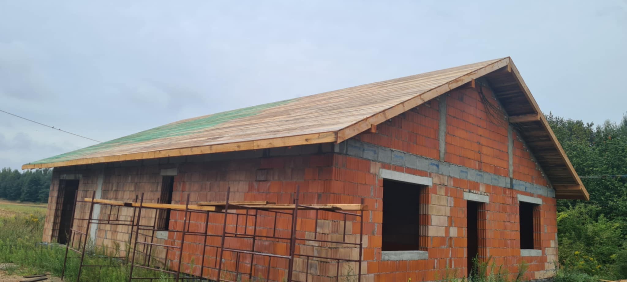 Surowy stan domu z czerwonej cegły z widoczną konstrukcją dachu z drewnianych łat. Częściowo zielona papa na dachu. Okna bez szyb. Otoczenie: trawa, drzewa.