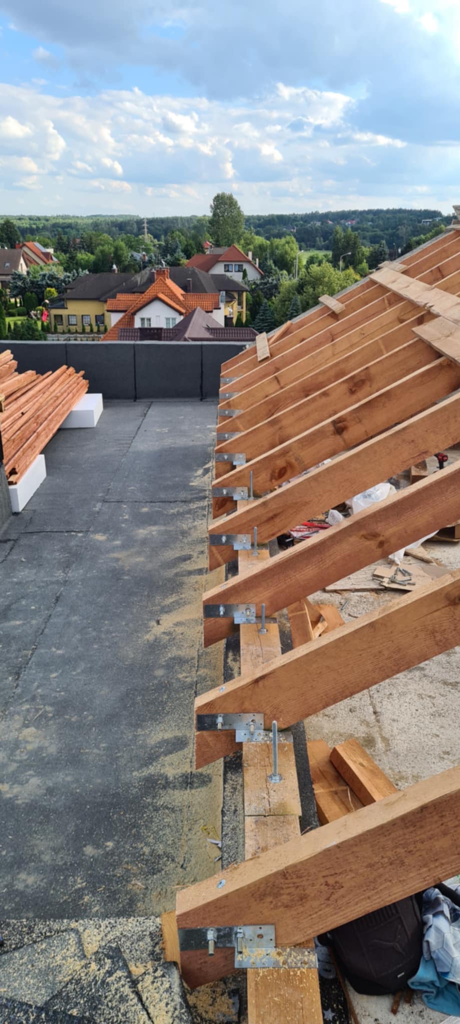 Wysoki kąt: drewniana więźba dachowa w trakcie montażu na płaskim dachu z widokiem na zielone tereny i zabudowę mieszkalną w tle, elementy mocujące widoczne.