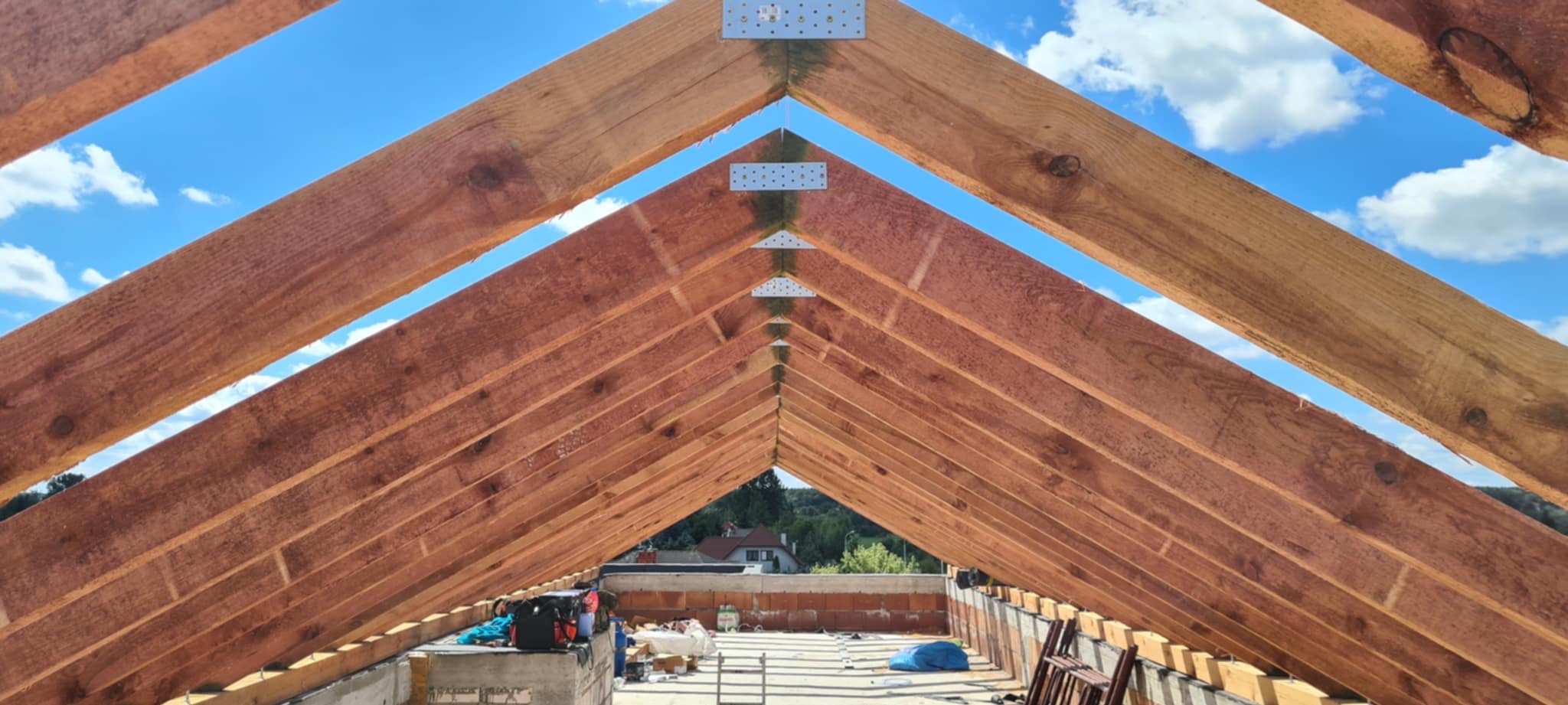 Wnętrze konstrukcji dachu z drewnianą więźbą, widok z dołu na niebieskie niebo. Metalowe łączniki wzmacniają konstrukcję. W tle widoczne narzędzia i materiały budowlane.