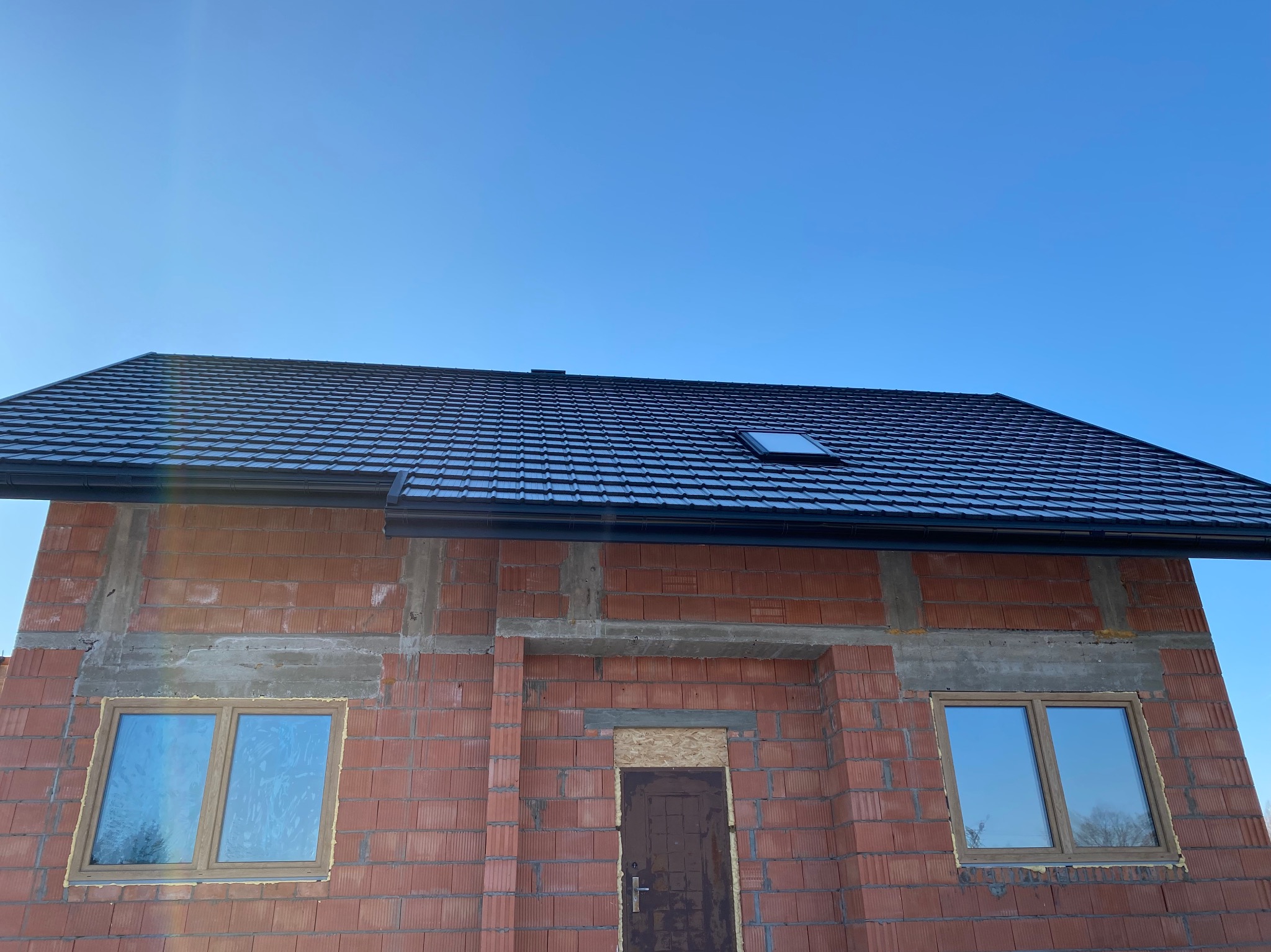 Dom w trakcie budowy z czarnym, błyszczącym dachem z dachówki modułowej i oknem dachowym. Widoczna surowa cegła i nieotynkowane ściany. Błękitne niebo w tle.