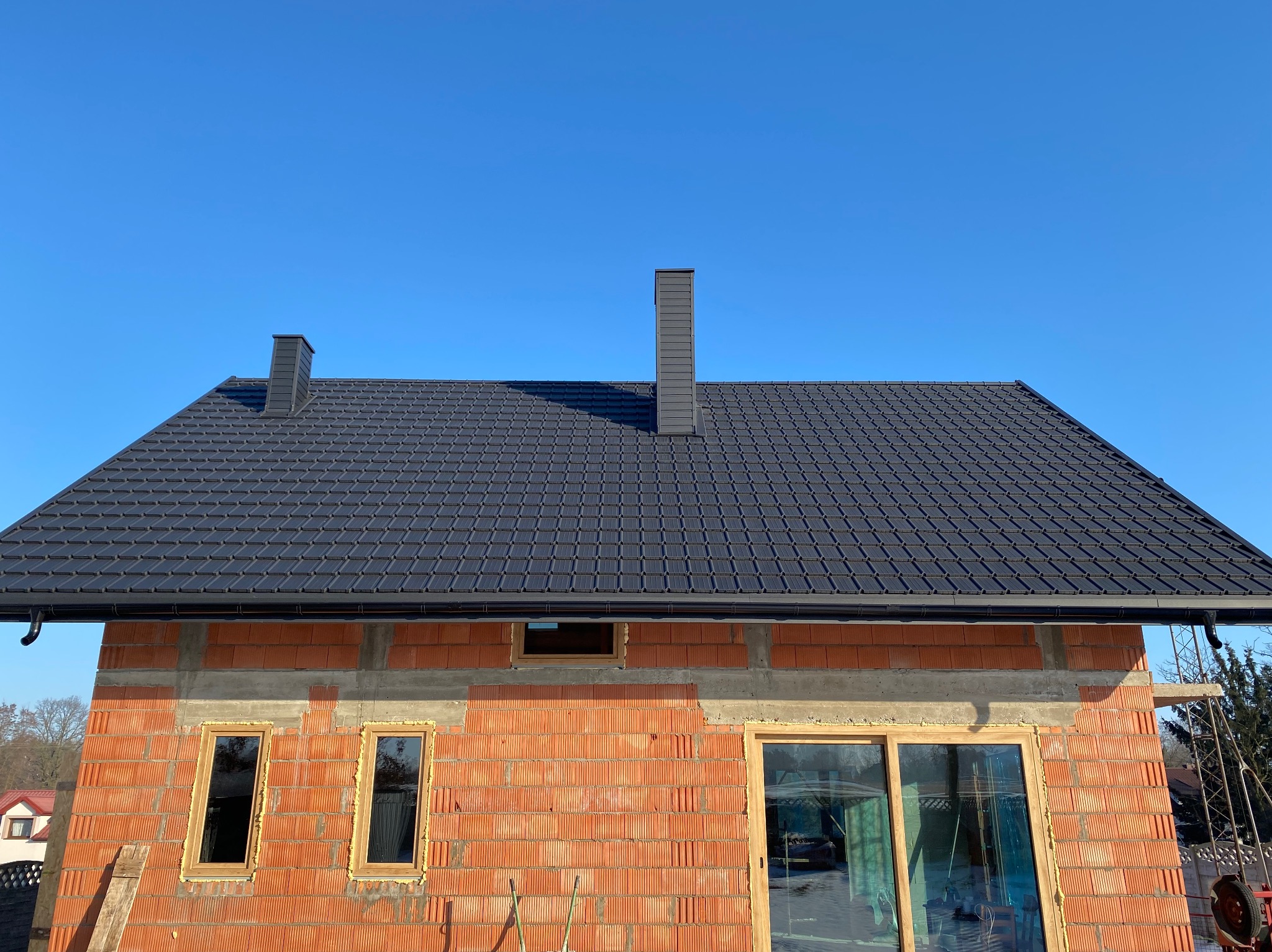 Dom w trakcie budowy z czarnym dachem z dachówki ceramicznej i dwoma kominami na tle błękitnego nieba. Widoczne okna i drzwi w stanie surowym.
