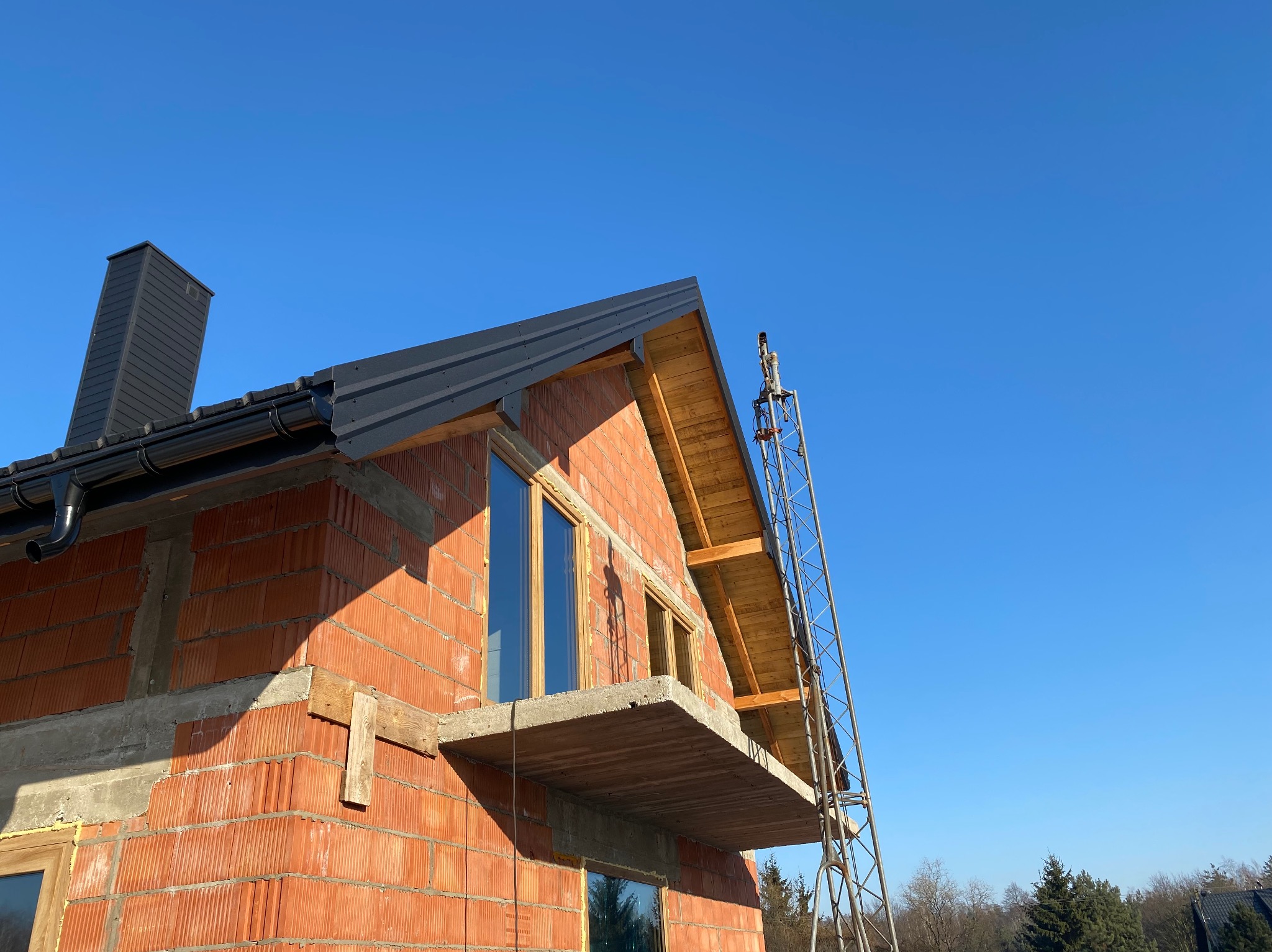 Budowa domu z czerwonej cegły w Dmosinie, widok z dołu na częściowo zadaszony budynek z oknami i rusztowaniem na tle błękitnego nieba. Etap prac dekarskich.