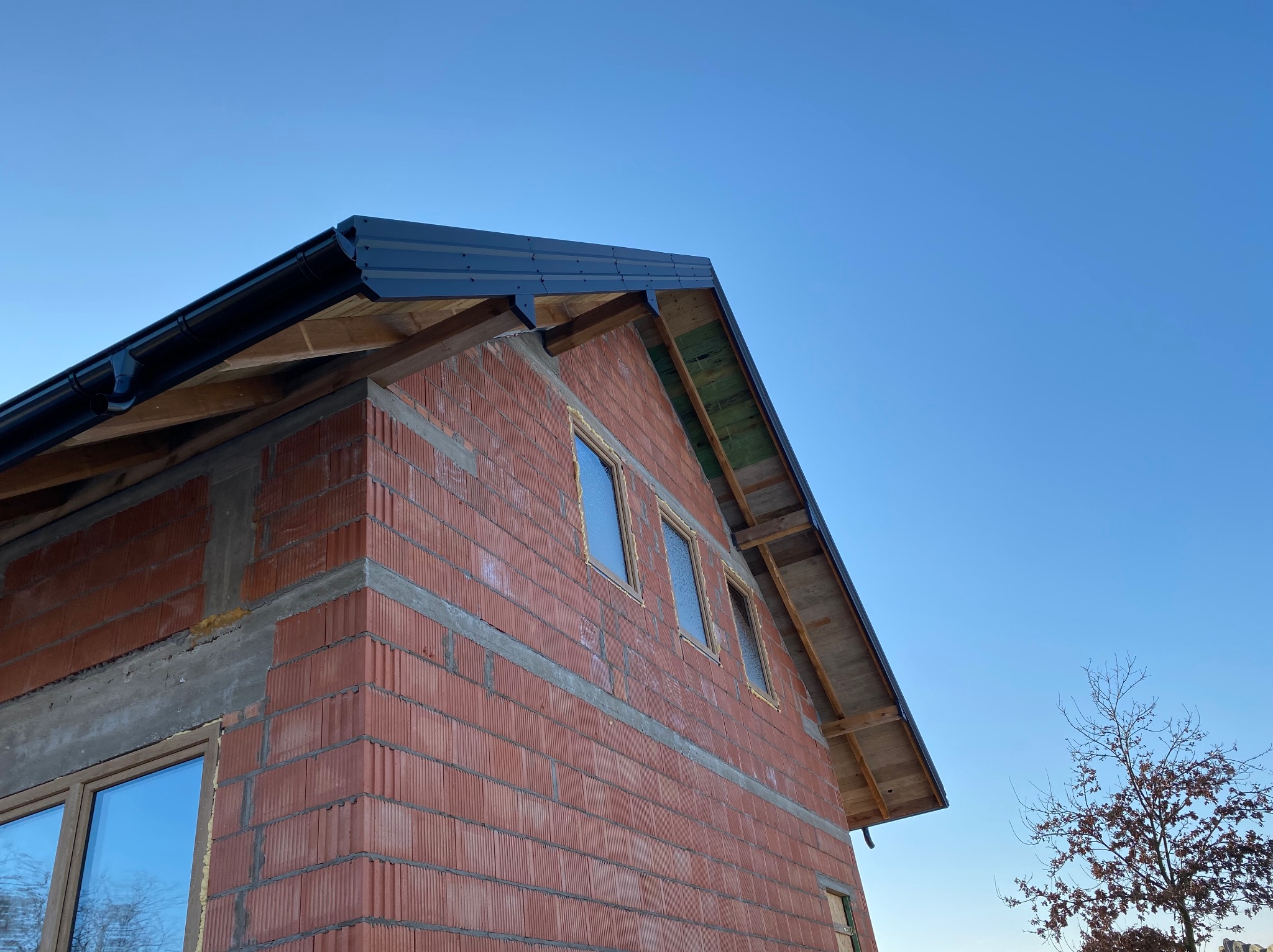 Surowy budynek z czerwonej cegły z zamontowaną czarną rynną i częściowo pokrytym dachem, widoczne drewniane elementy konstrukcji dachu na tle błękitnego nieba.