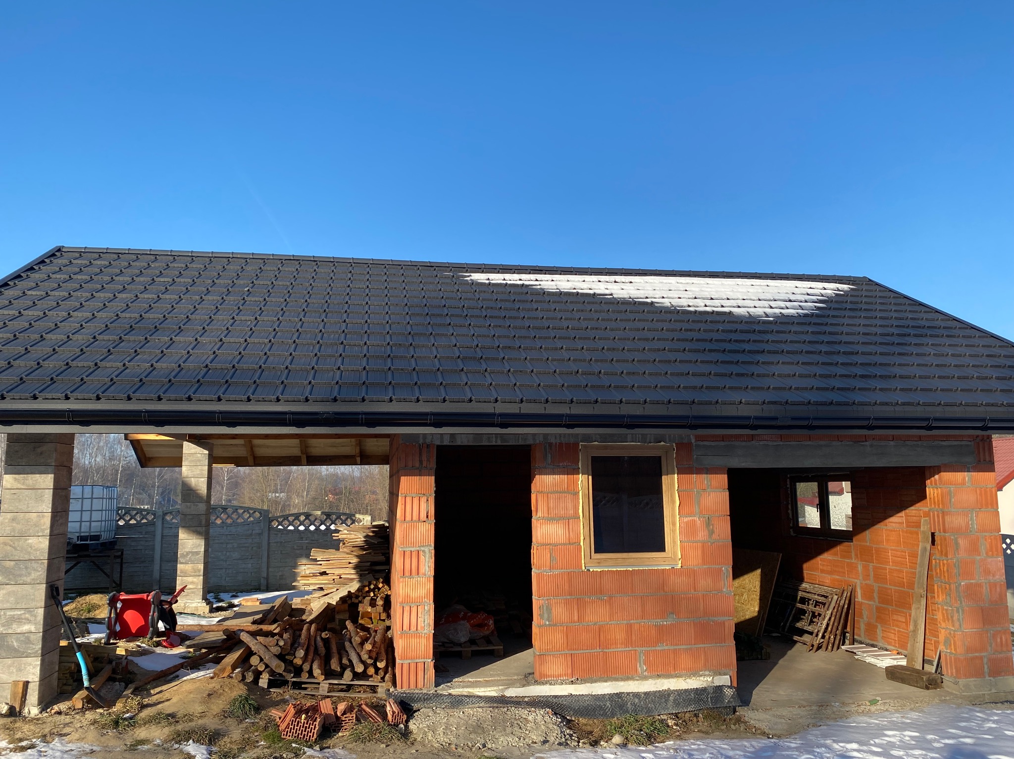 Nowy, czarny dach z blachodachówki na budynku w trakcie budowy, częściowo pokryty śniegiem. Widoczne ceglane ściany i drewniane elementy konstrukcyjne.