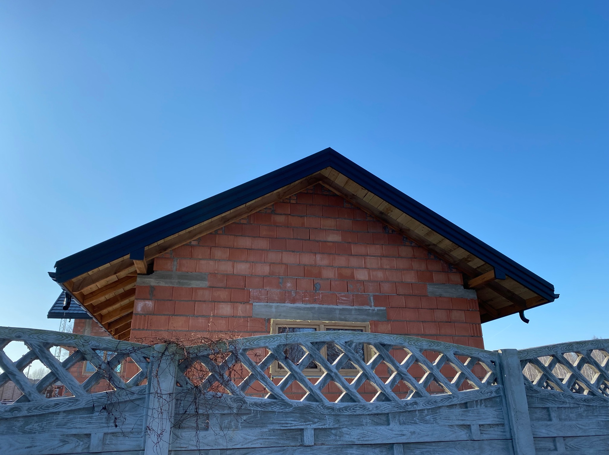 Surowy budynek z czerwonej cegły z czarnym dachem, widoczny zza betonowego ażurowego ogrodzenia na tle błękitnego nieba. Widoczne elementy konstrukcji dachu.