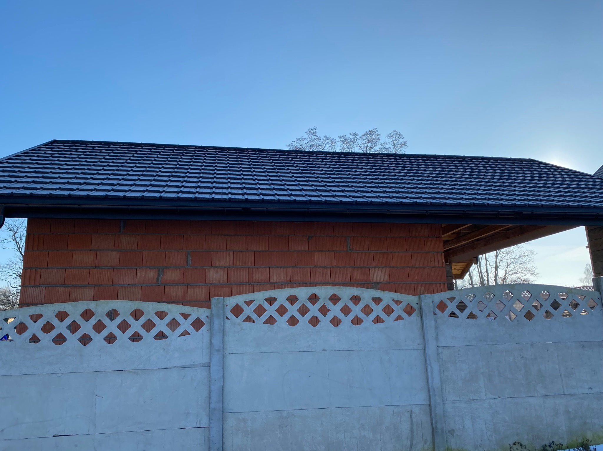 Nowy, czarny dach z dachówki ceramicznej na budynku z czerwonej cegły, widziany zza betonowego ogrodzenia w słoneczny dzień. Widoczne rynny i fragment konstrukcji dachu.