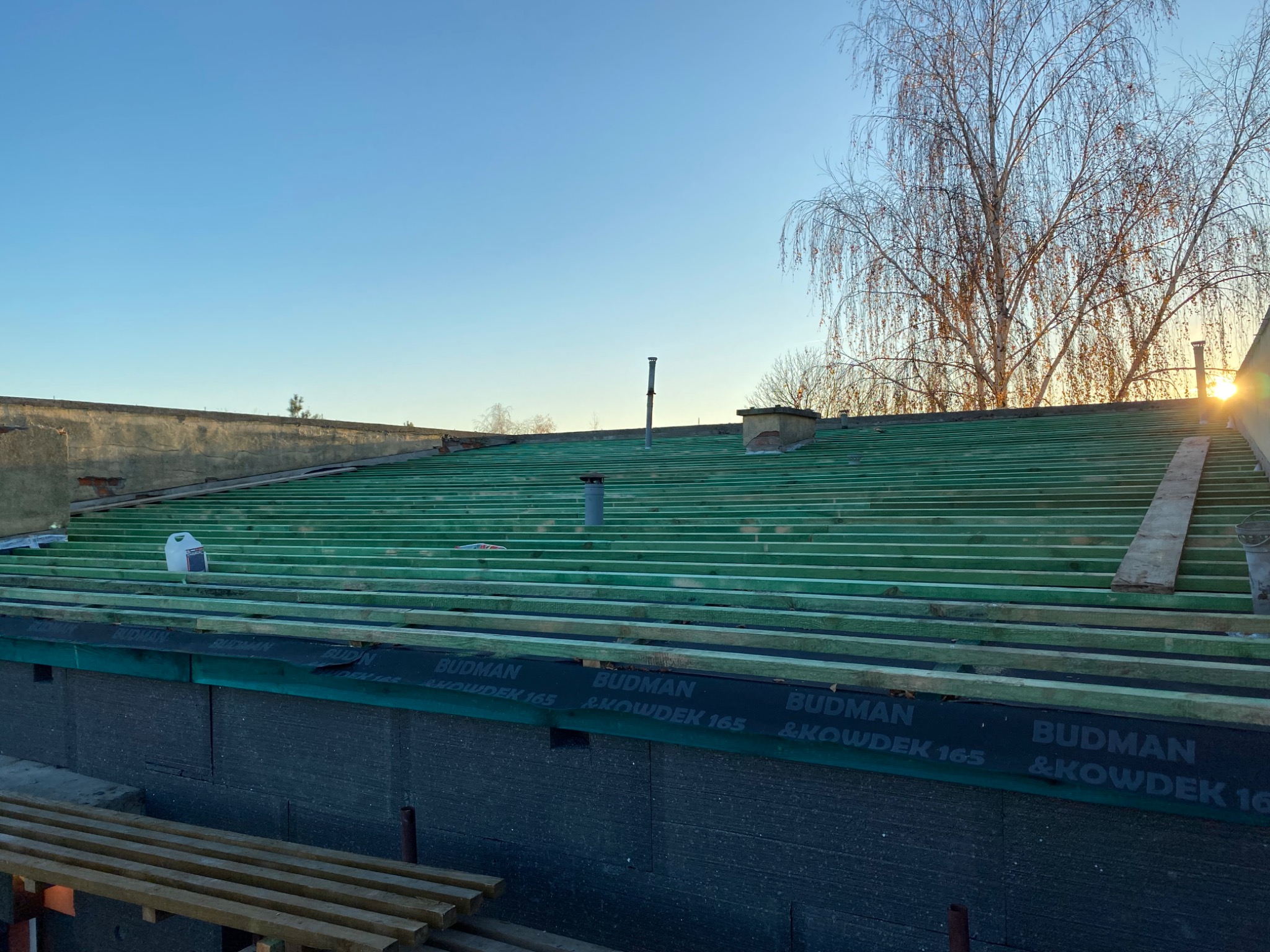 Zielone łaty na dachu z widoczną membraną Budman & Kowdek 165, komin i drzewo w tle, wczesny etap prac dekarskich w słoneczny dzień.