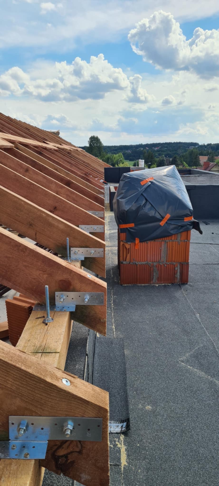 Wiązar dachowy z drewnianymi belkami i metalowymi łącznikami na tle pokrycia dachowego z papy. Mur z cegieł zabezpieczony folią. Widok na okolicę i błękitne niebo.