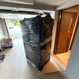 Zenon Światłoch - Lodówka owinięta czarną folią stretch, przygotowana do transportu. Widoczne otwarte drzwi garażowe i fragment podjazdu.
