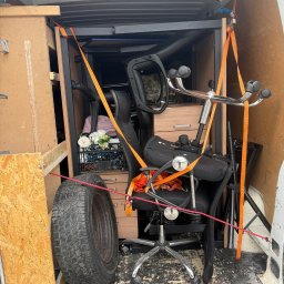 Zenon Światłoch - Wnętrze busa załadowane meblami biurowymi, kwiatami w skrzynce i oponą, zabezpieczone pasami transportowymi. Widoczne elementy mocujące.