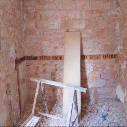 Turbo Lokalnie - Pomieszczenie z odsłoniętymi ceglanymi ścianami i częściowo usuniętym tynkiem, widoczne korytko na instalacje. Na podłodze gruz, a w tle drewniana deska i koziołek budowlany.