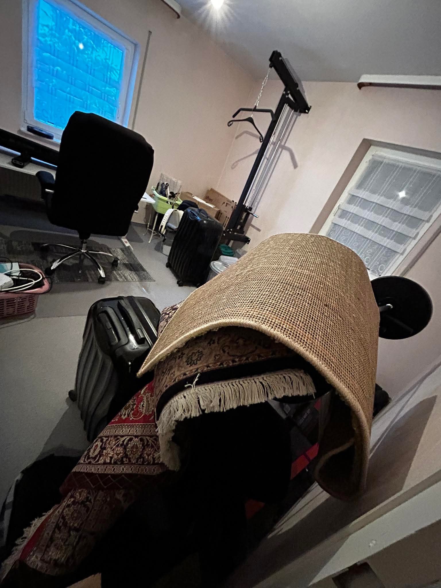 Pokój w trakcie przygotowań do transportu: walizki, zwinięte dywany, krzesło biurowe i sprzęt do ćwiczeń. Widok na okno.