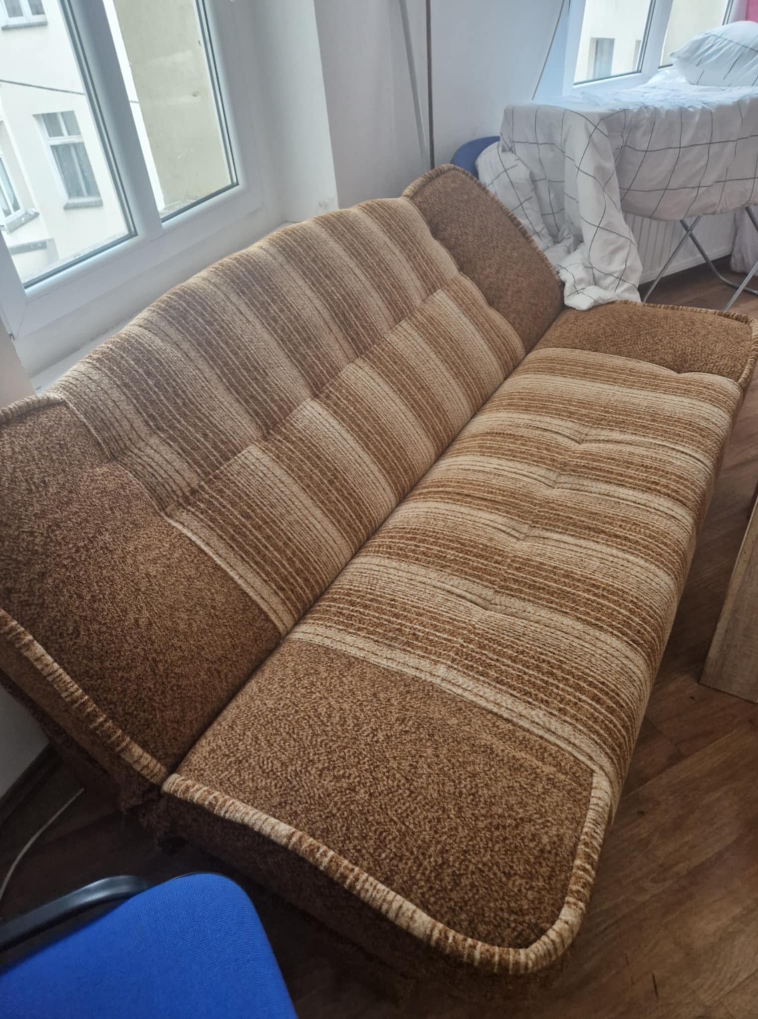 Rozłożona sofa w brązowe pasy stoi w pokoju przy oknie. Obok widoczny jest rozkładany suszak na pranie i fragment krzesła.
