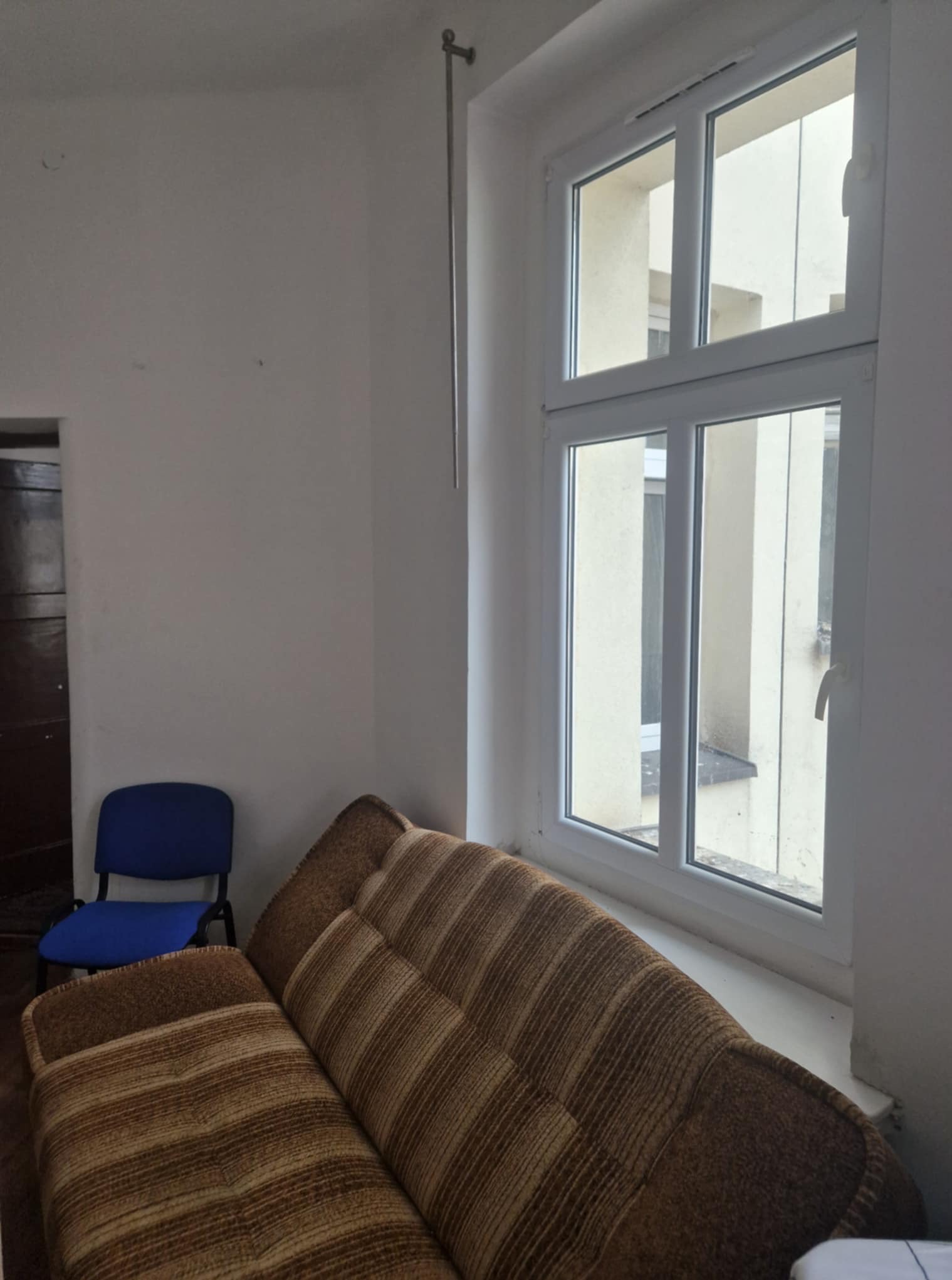 Pokój z brązową kanapą w paski i niebieskim krzesłem, oświetlony przez okno z białymi ramami. Widoczne ślady użytkowania i brak dekoracji.
