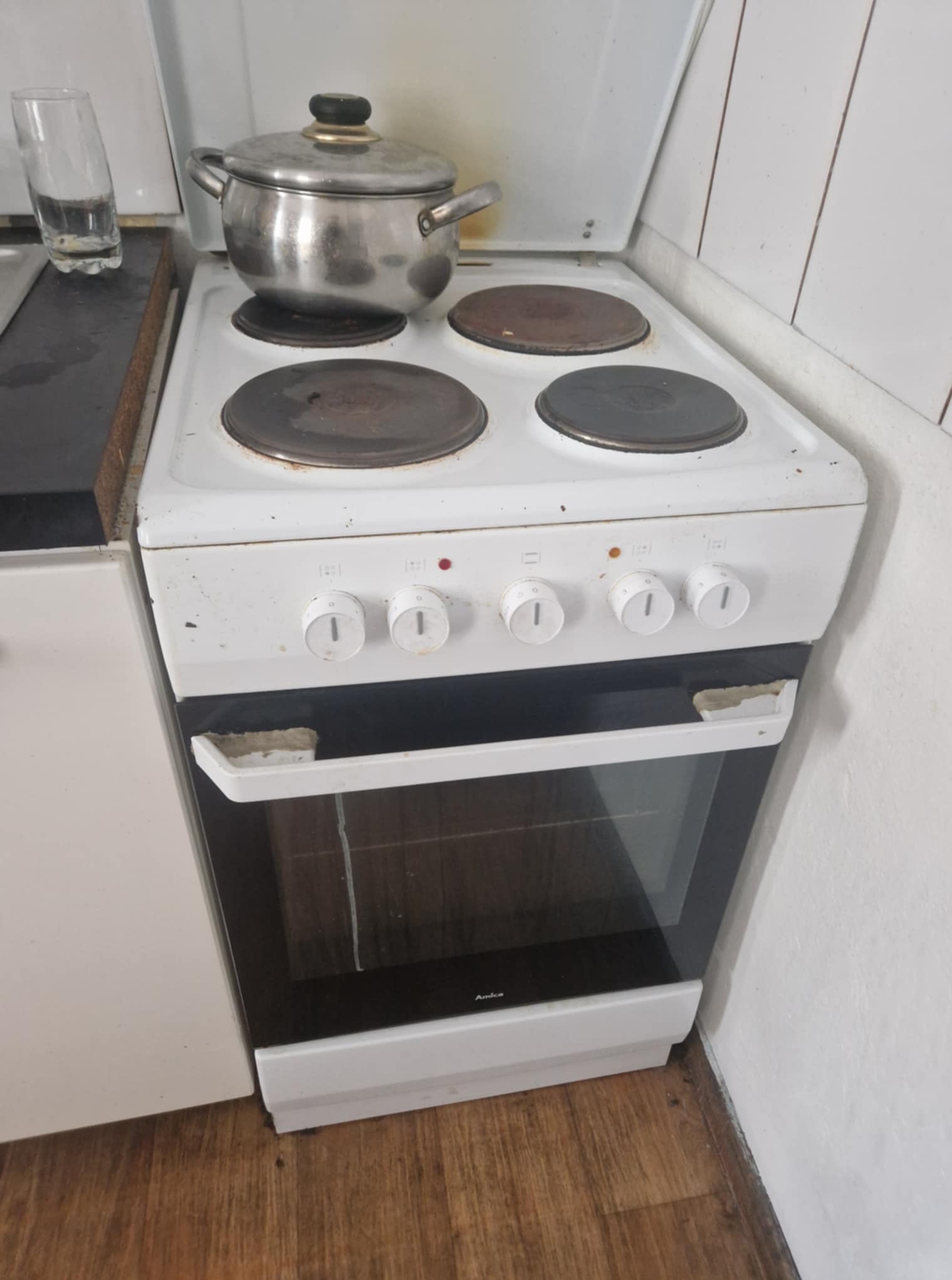 Biała, używana kuchenka elektryczna Amica z czterema palnikami, na jednym z nich stoi metalowy garnek. Widoczne zabrudzenia i uszkodzenia na obudowie.