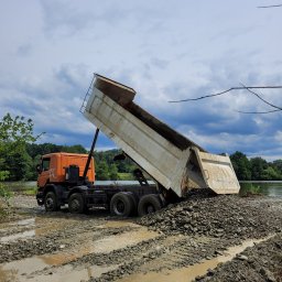 TMG - Pomarańczowa wywrotka z podniesioną skrzynią wysypuje gruz nad brzegiem zbiornika wodnego. Błotnista droga, pochmurne niebo.