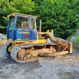 Żółto-niebieski spychacz Hanomag D 600 D na żwirowym podłożu, z zielonymi drzewami w tle. Maszyna gotowa do pracy przy niwelacji terenu lub wykopach pod fundamenty.