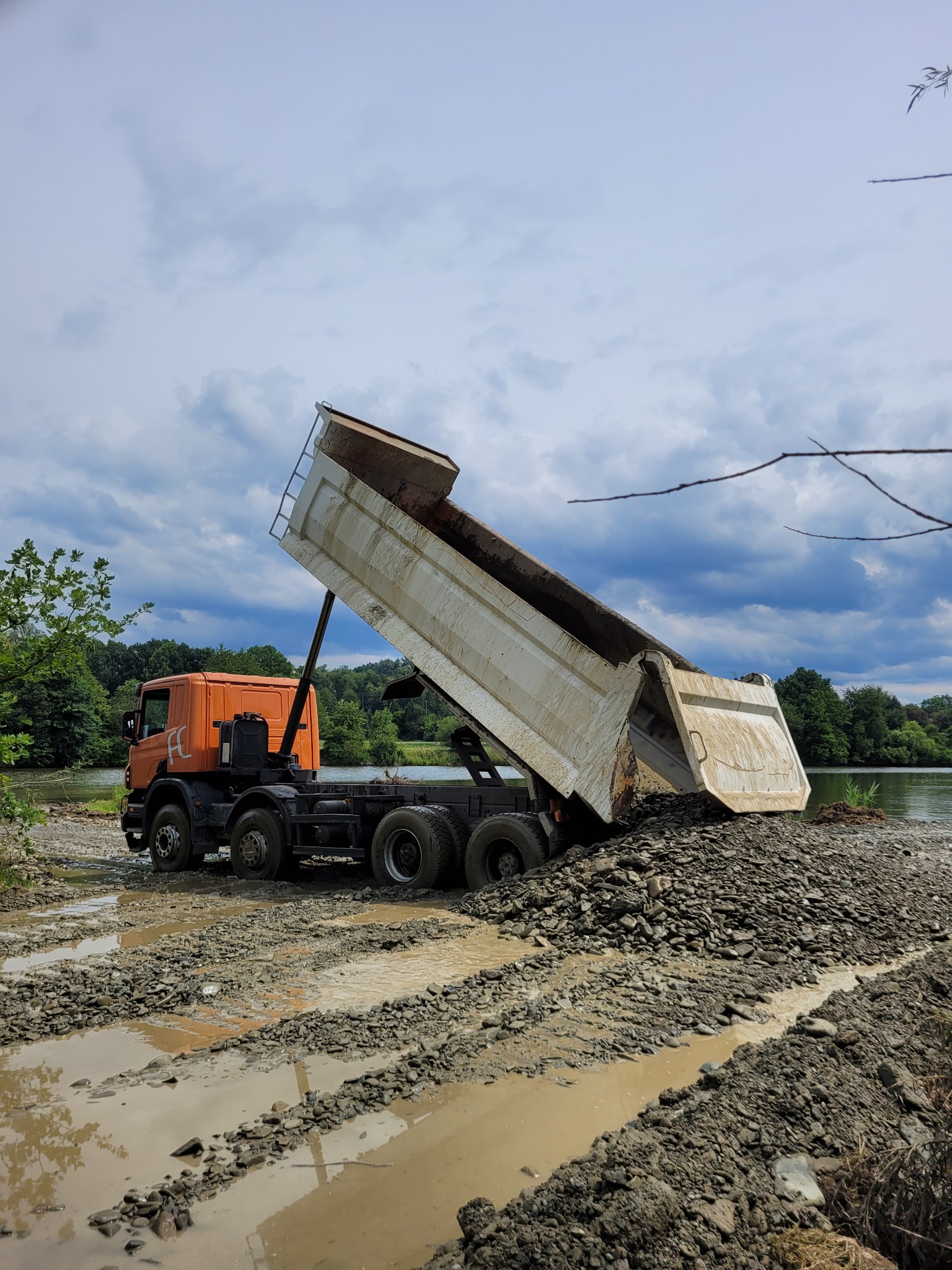 Pomarańczowa wywrotka z podniesioną skrzynią wysypuje gruz nad brzegiem zbiornika wodnego. Błotnista droga, pochmurne niebo.