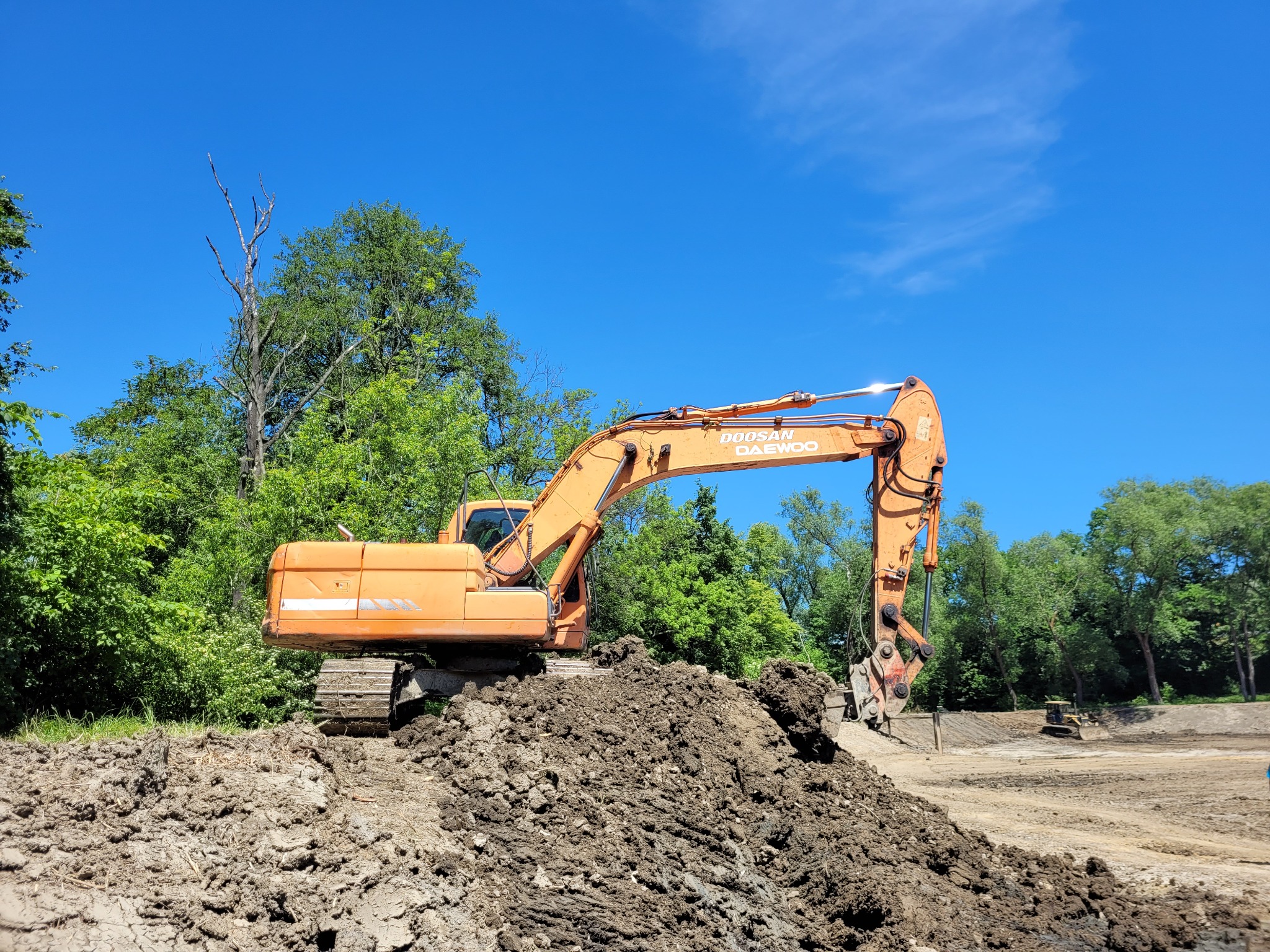 Pomarańczowa koparka Doosan Daewoo na hałdzie ziemi, w tle zieleń drzew i błękit nieba. Widoczny kolejny sprzęt budowlany w oddali.