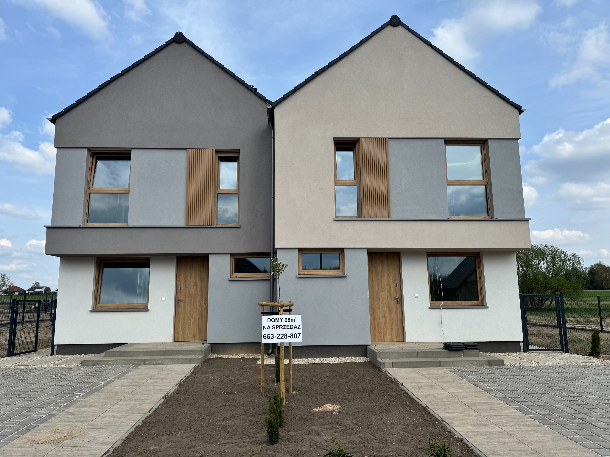 Dwa segmenty bliźniacze w odcieniach szarości i beżu, z drewnianymi akcentami i oknami. Na trawniku tablica z informacją o sprzedaży. Widok z przodu, błękitne niebo.