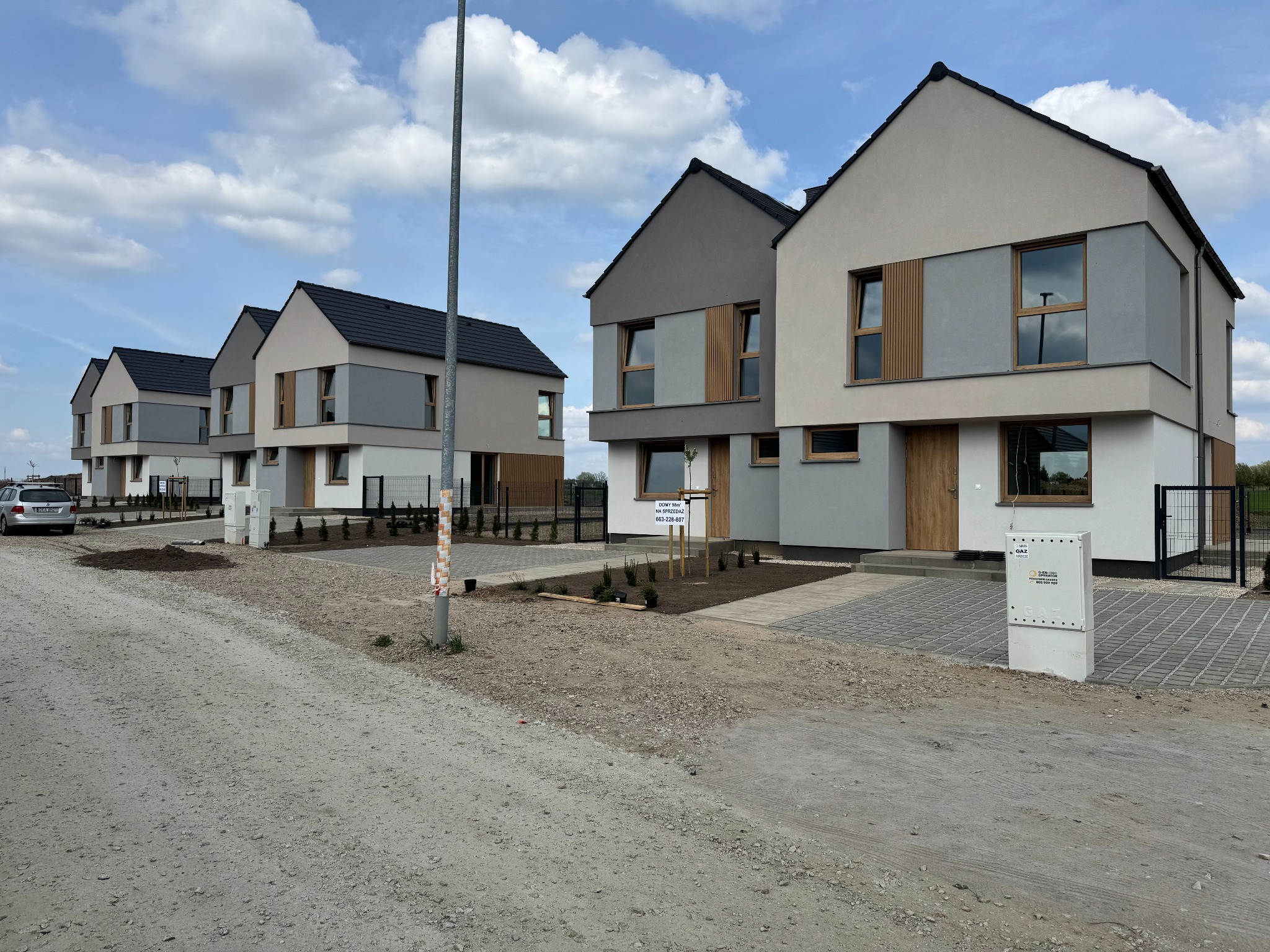 Nowoczesne domy szeregowe z elewacją w odcieniach szarości i beżu, drewniane akcenty, kostka brukowa na podjeździe, błękitne niebo w tle.