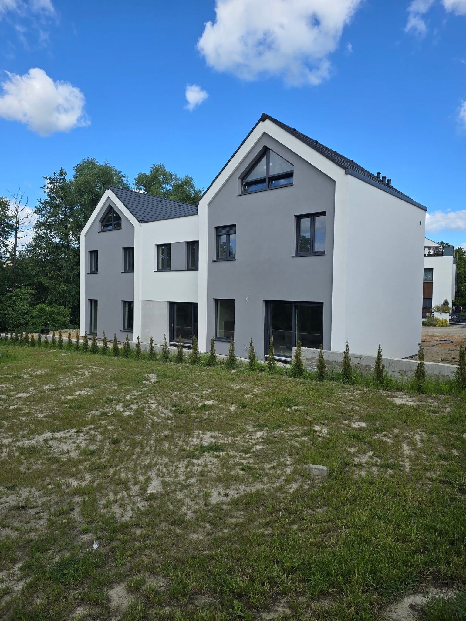 Dwa nowoczesne domy w odcieniach szarości i bieli, z ciemnymi dachami i oknami, otoczone młodymi drzewkami. Błękitne niebo z chmurami w tle.