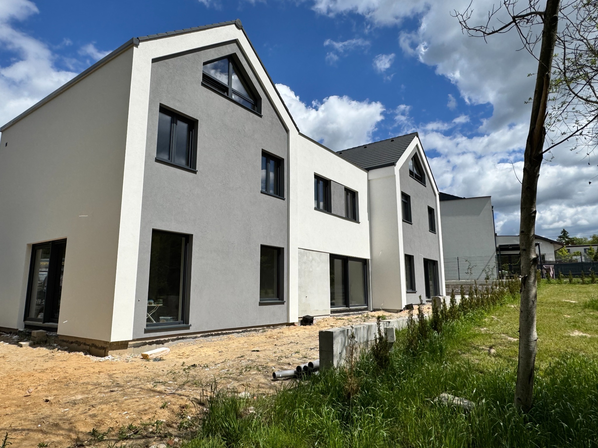 Nowoczesny szeregowiec z elewacją w bieli i szarości, czarne okna, trawnik w budowie, błękitne niebo. Minimalistyczny design.