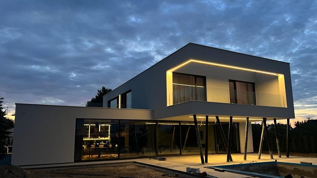 Nowoczesny dom z oświetloną elewacją i balkonem, sfotografowany o zmierzchu, z widocznym basenem w budowie. Minimalistyczny design.