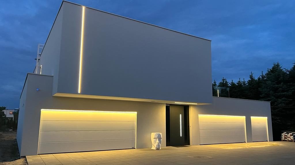 Nowoczesny dom z płaskim dachem oświetlony ledami w pionowych liniach i nad bramami garażowymi, minimalistyczna bryła budynku w szarym kolorze.