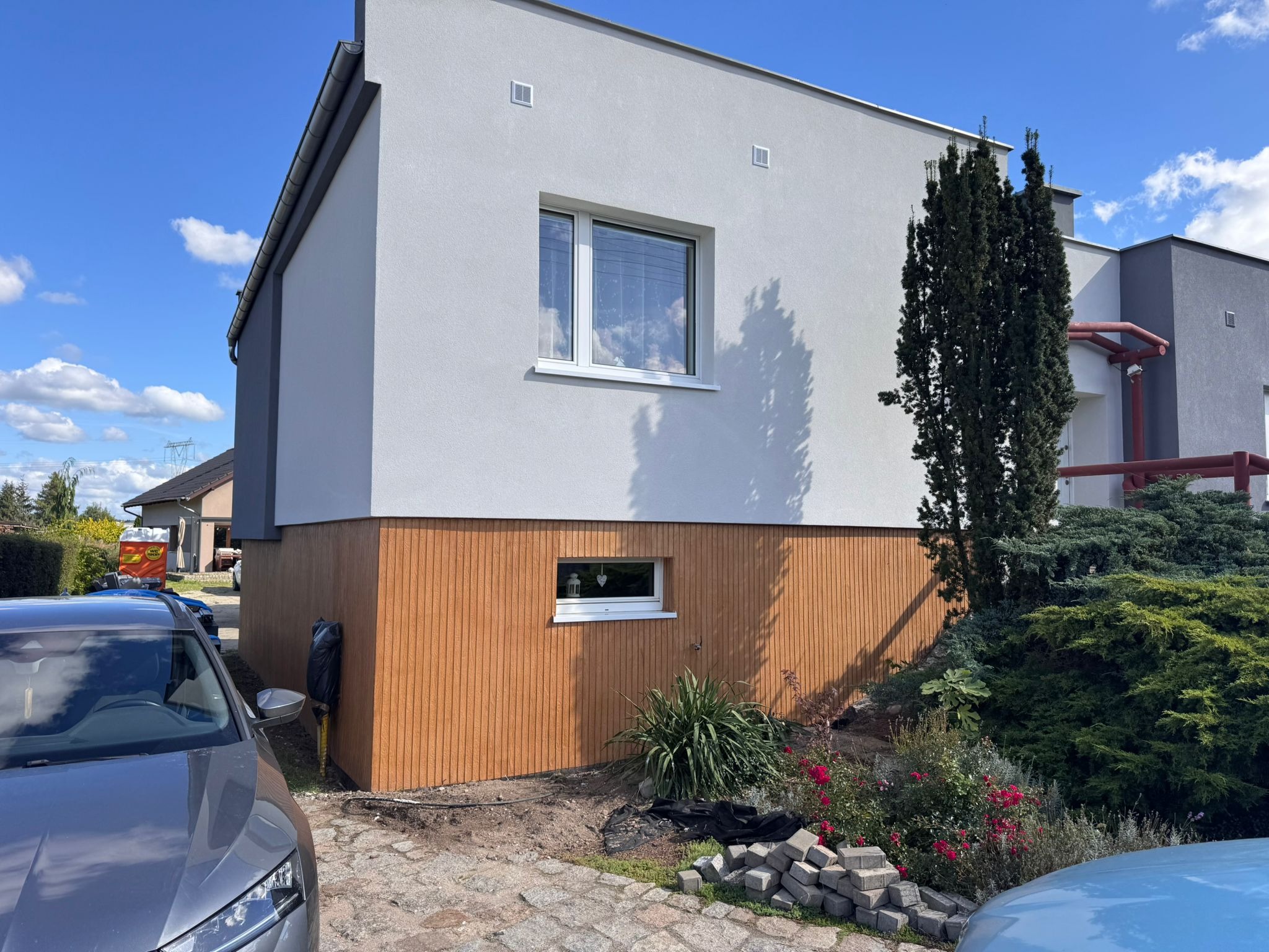 Nowoczesny dom z jasnoszarą elewacją i drewnopodobną okładziną na dole, okna z białymi ramami, kostka brukowa i roślinność w otoczeniu. Błękitne niebo w tle.