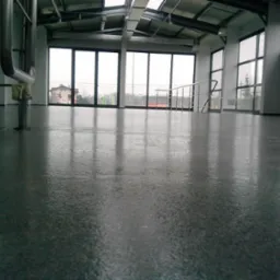 Szara, błyszcząca posadzka w hali przemysłowej z widocznymi metalowymi rurami, oknami i elementami konstrukcji dachu.