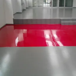 Częściowo wykończona podłoga w pomieszczeniu, z fragmentami w kolorze czerwonym i szarym, z widocznymi refleksami światła i schodami prowadzącymi do wyższego poziomu.