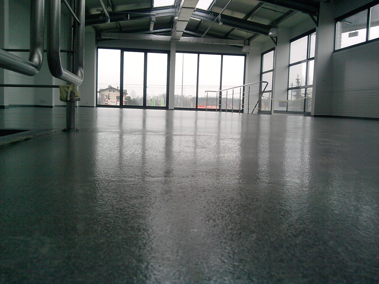 Szara, błyszcząca posadzka w hali przemysłowej z widocznymi metalowymi rurami, oknami i elementami konstrukcji dachu.