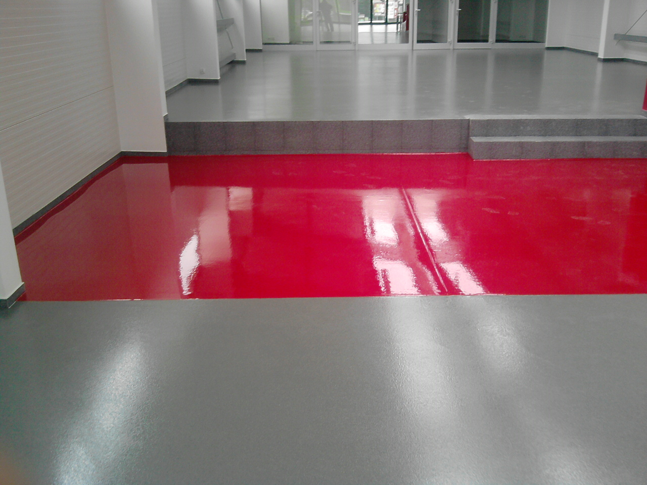 Częściowo wykończona podłoga w pomieszczeniu, z fragmentami w kolorze czerwonym i szarym, z widocznymi refleksami światła i schodami prowadzącymi do wyższego poziomu.