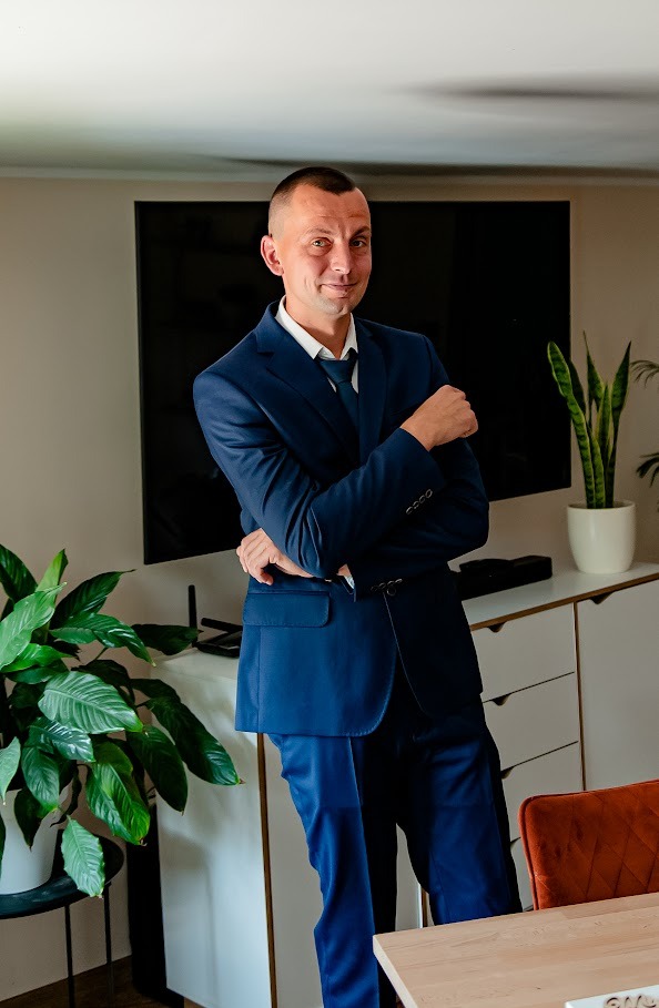 Mężczyzna w granatowym garniturze, oparty o blat. W tle biała komoda, telewizor i rośliny doniczkowe. Uśmiechnięty, pewny siebie, w jasnym pomieszczeniu.