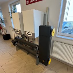 MULTIHEAT PATRYK SZUBIŃSKI - Dwa białe piece gazowe Ariston z czarnym zasobnikiem ciepłej wody, połączone rurami. Widoczny grzejnik i roślina doniczkowa. Jasne wnętrze, płytki podłogowe.