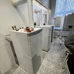 MULTIHEAT PATRYK SZUBIŃSKI - Białe, wiszące piece gazowe Vaillant z podłączonymi rurami, narzędzia na górze. Widok na okno z żaluzjami. Szary wzór na podłodze. Częściowo widoczna osoba.