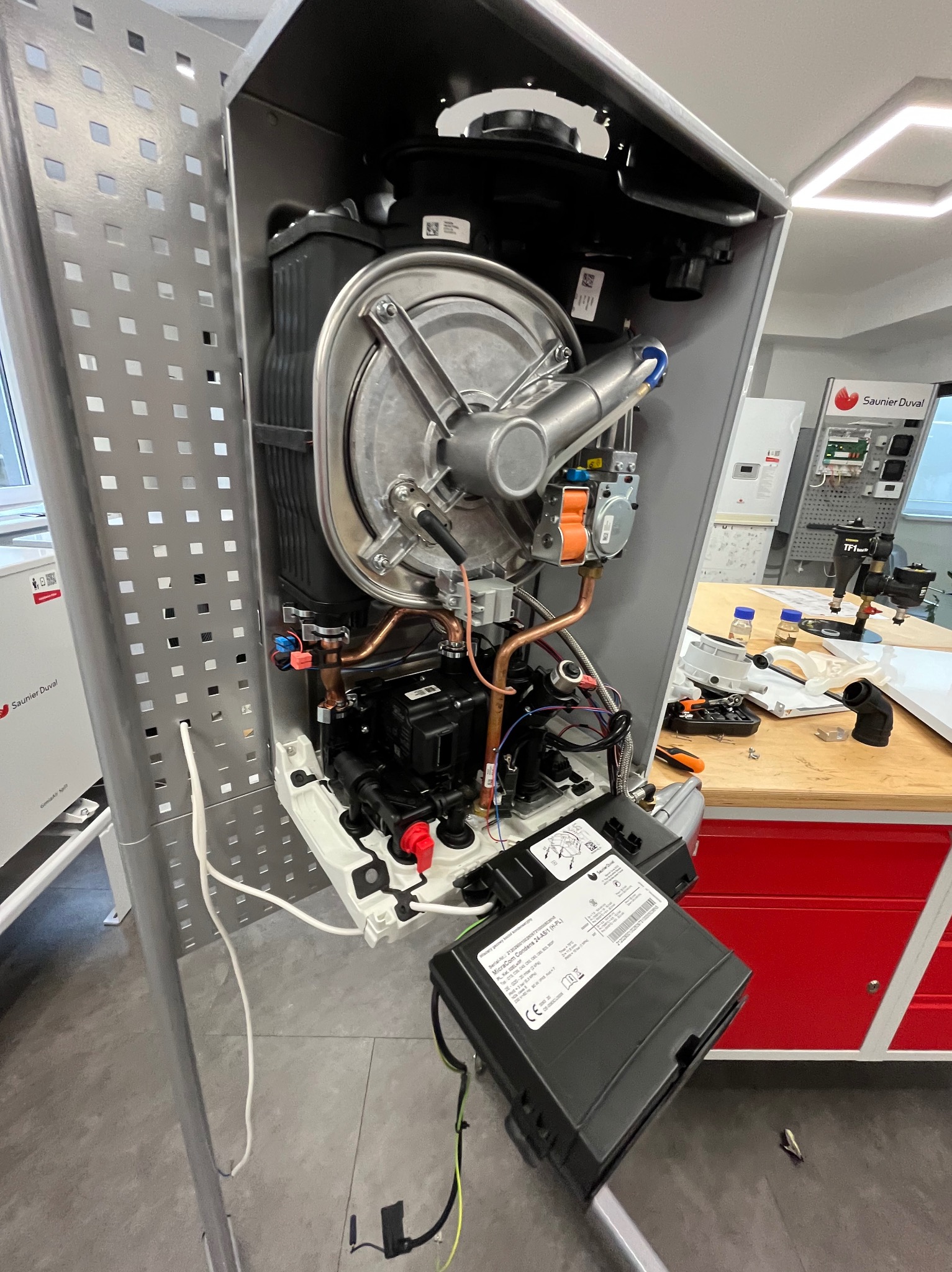 Otwarty piec gazowy marki Saunier Duval, widoczne elementy instalacji, miedziane rurki, czarna obudowa. Na drugim planie narzędzia i stół warsztatowy.